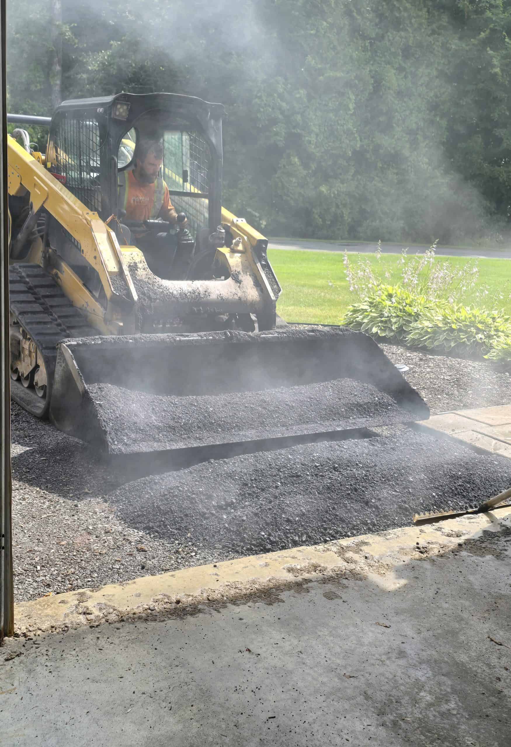 A construction worker operates a yellow compact track loader to dump hot asphalt onto a driveway, with steam rising heavily from the material.