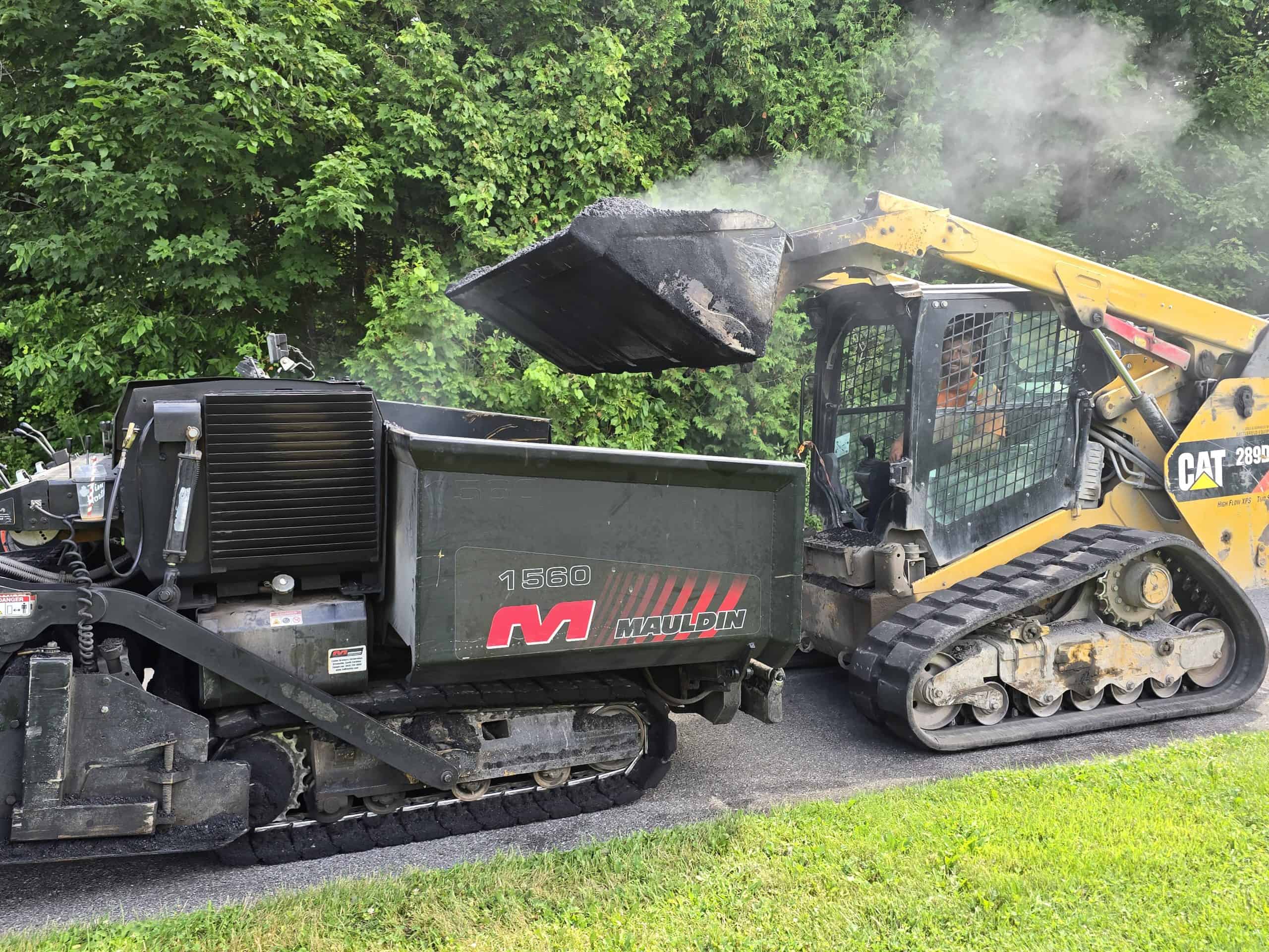 A CAT track loader dumps hot asphalt into the hopper of a Mauldin 1560 paver. Steam rises from the hot material during the paving operation.