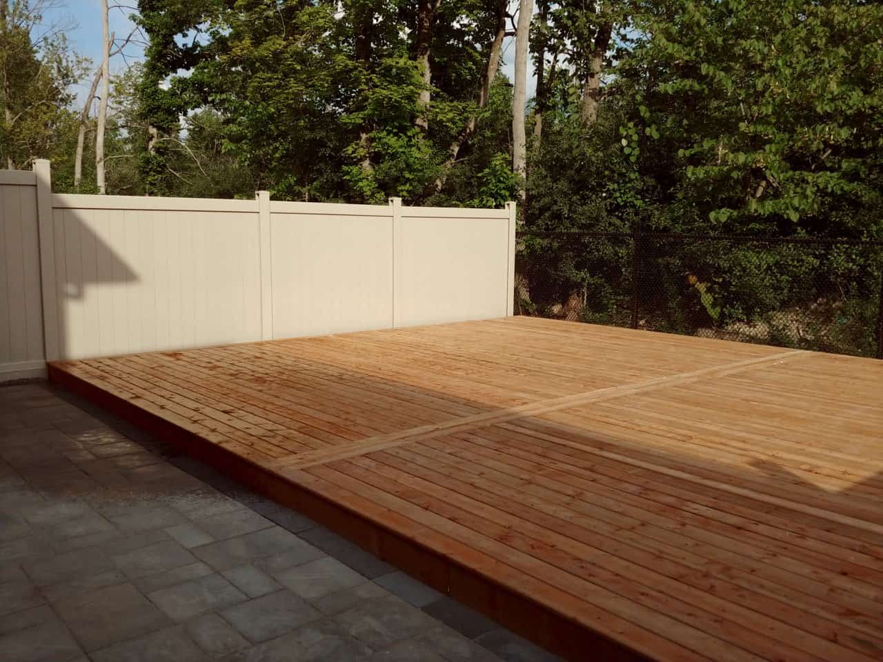 Low wood deck and vinyl fence bordering a paved patio. The clean, modern design provides a private outdoor living space next to trees.