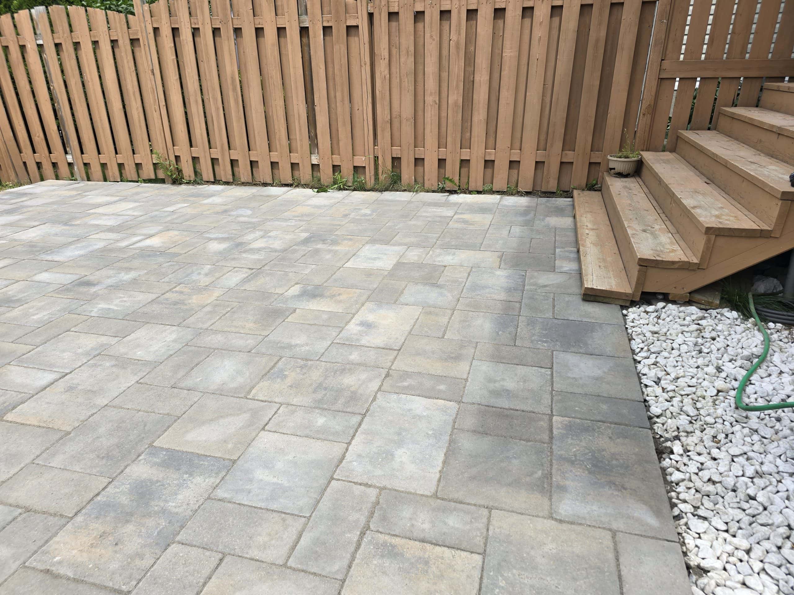 Newly finished residential patio featuring multi-toned paver hardscape. It meets a wooden fence and wooden deck stairs, with a clean border of white drainage stones.