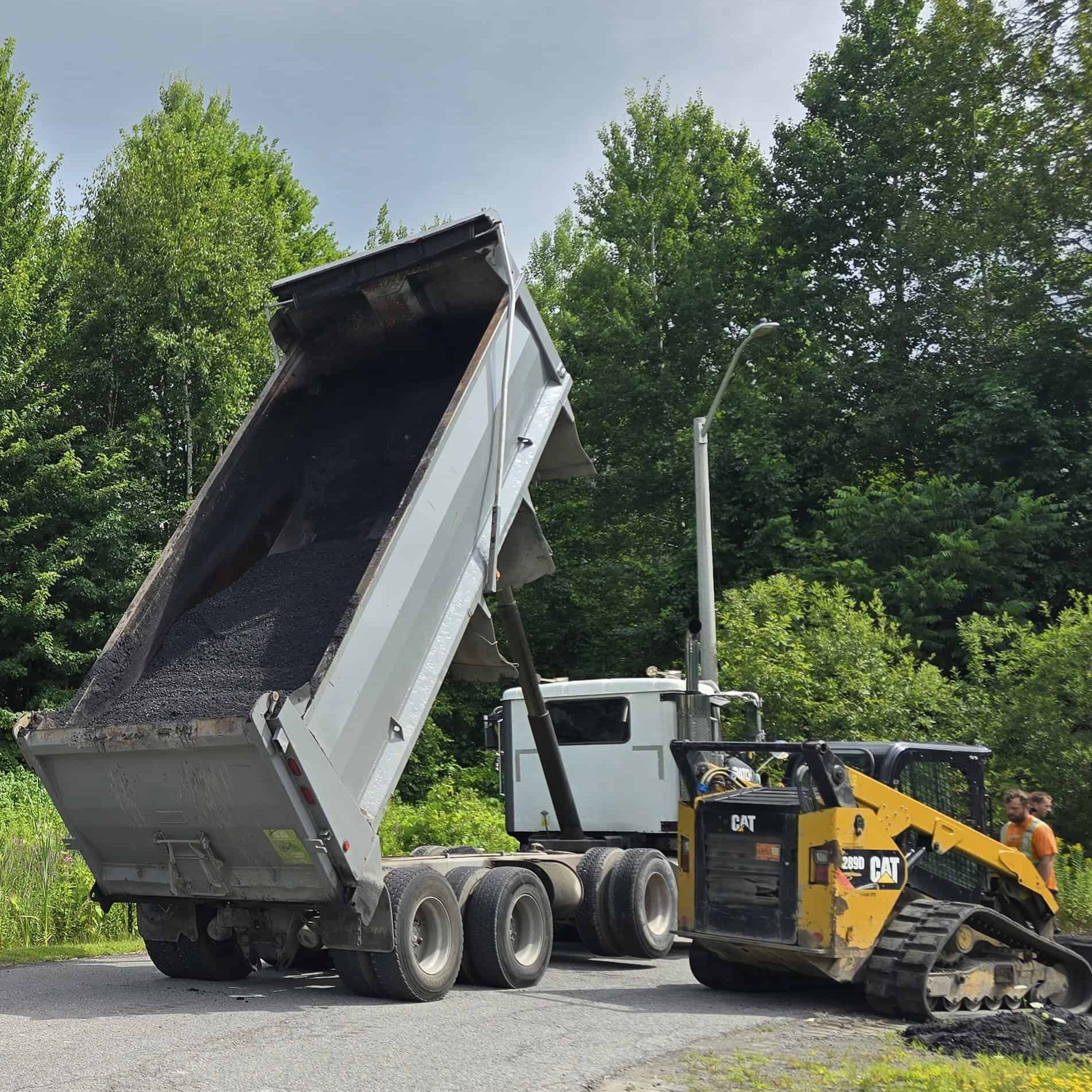Asphalt paving heavy equipment Heavy construction equipment for asphalt paving. A large dump truck unloads material next to a CAT skid steer loader on a road bordered by dense forest.