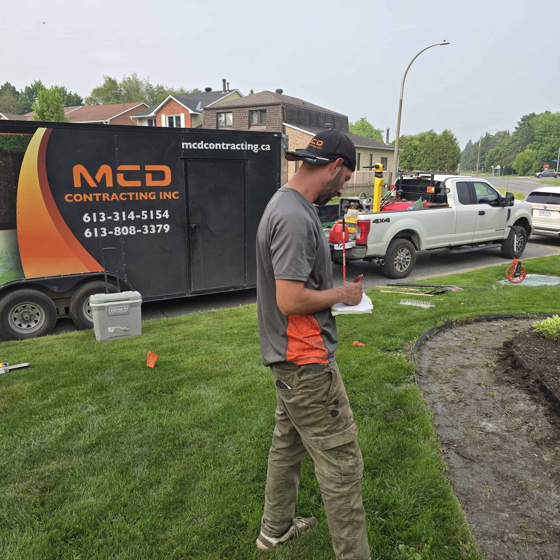 An MCD Contracting team member uses a laser level and takes notes on a project site, with the branded trailer nearby, preparing for high-quality landscaping or interlock work.