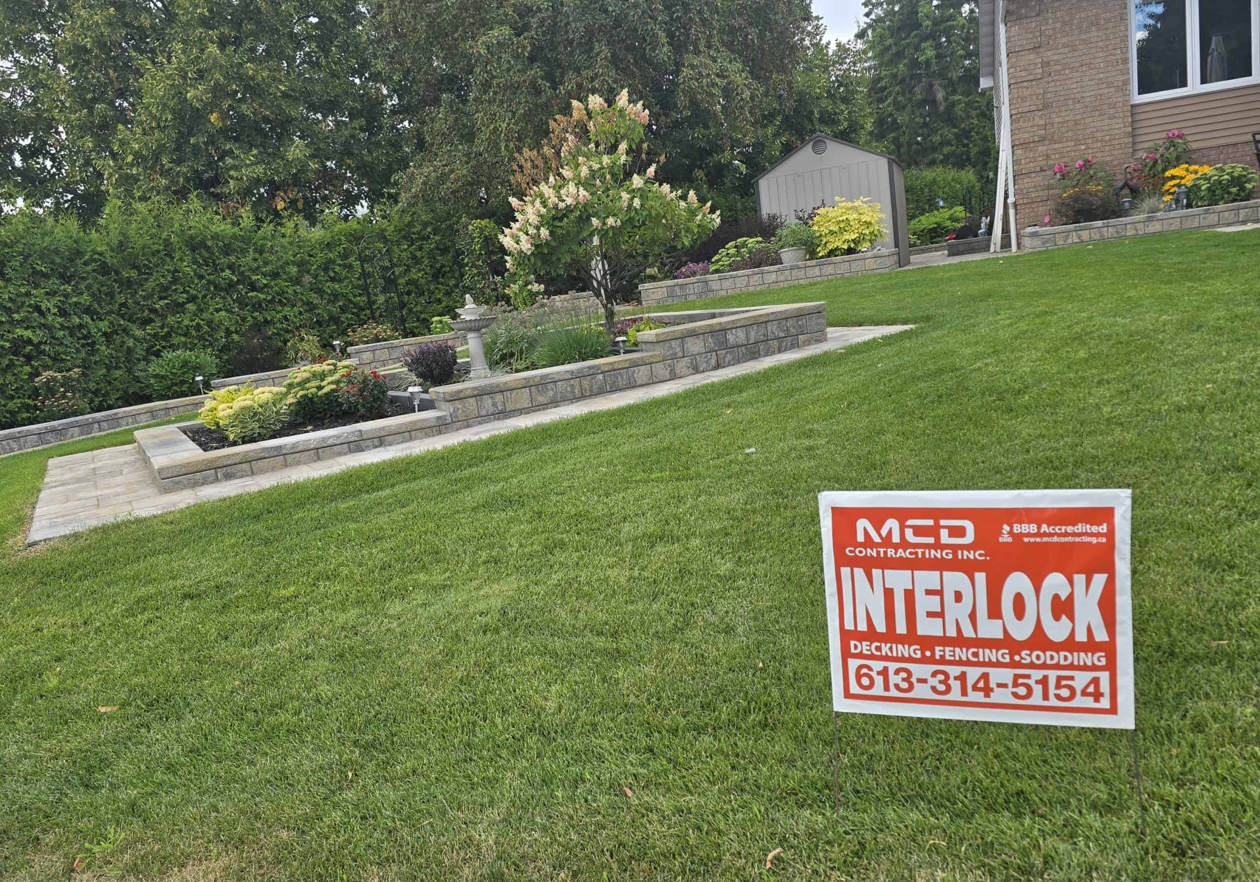 This image showcases a beautiful landscape design featuring tiered interlock retaining walls and garden beds on a sloping lawn, completed by MCD Contracting.
