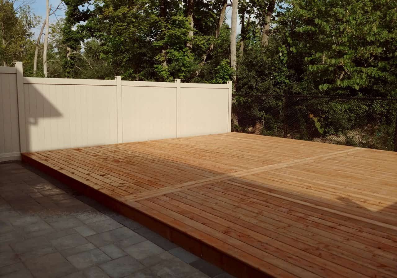Low wood deck and vinyl fence bordering a paved patio. The clean, modern design provides a private outdoor living space next to trees.