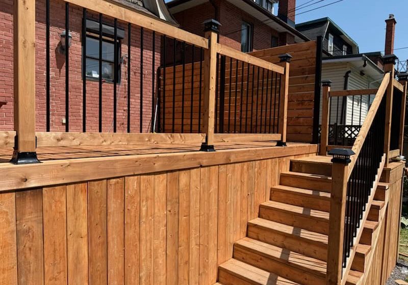 Stone stepping stone gravel path A large wooden deck with stairs features a black railing system and wood skirting, next to a red brick house.